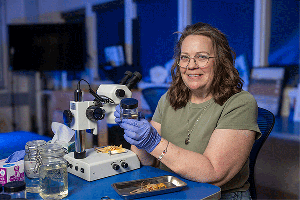 Middle-aged white female with shoulder-length brown hair and glasses sits at a table in a lab setting and holds in her hands a jar that contains a crayfish. Another crayfish is visible on a tray underneath a microscope. 